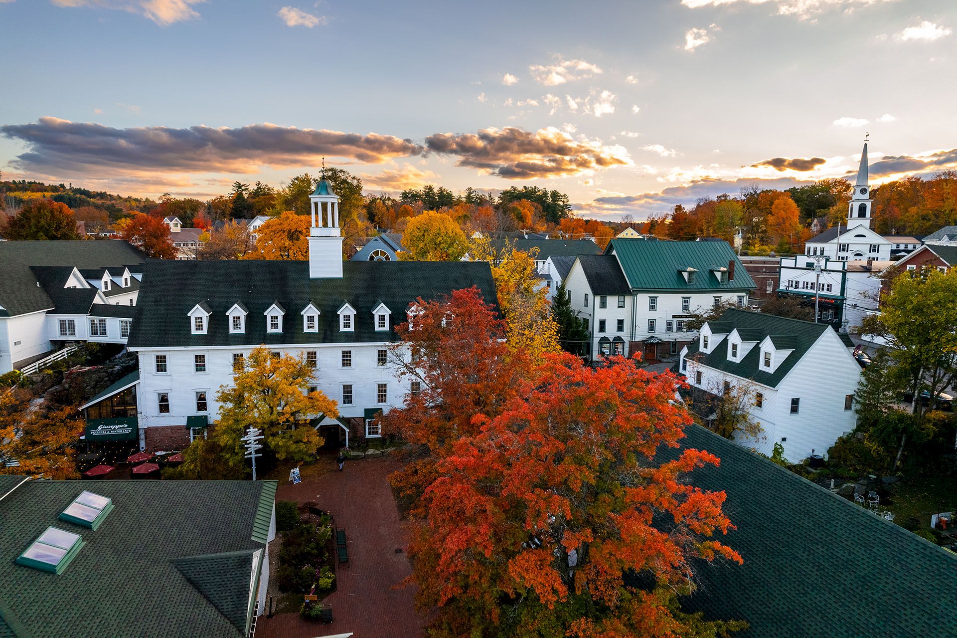 An aerial view of a white campus building with a steeple, surrounded by vivid autumn trees at sunset.