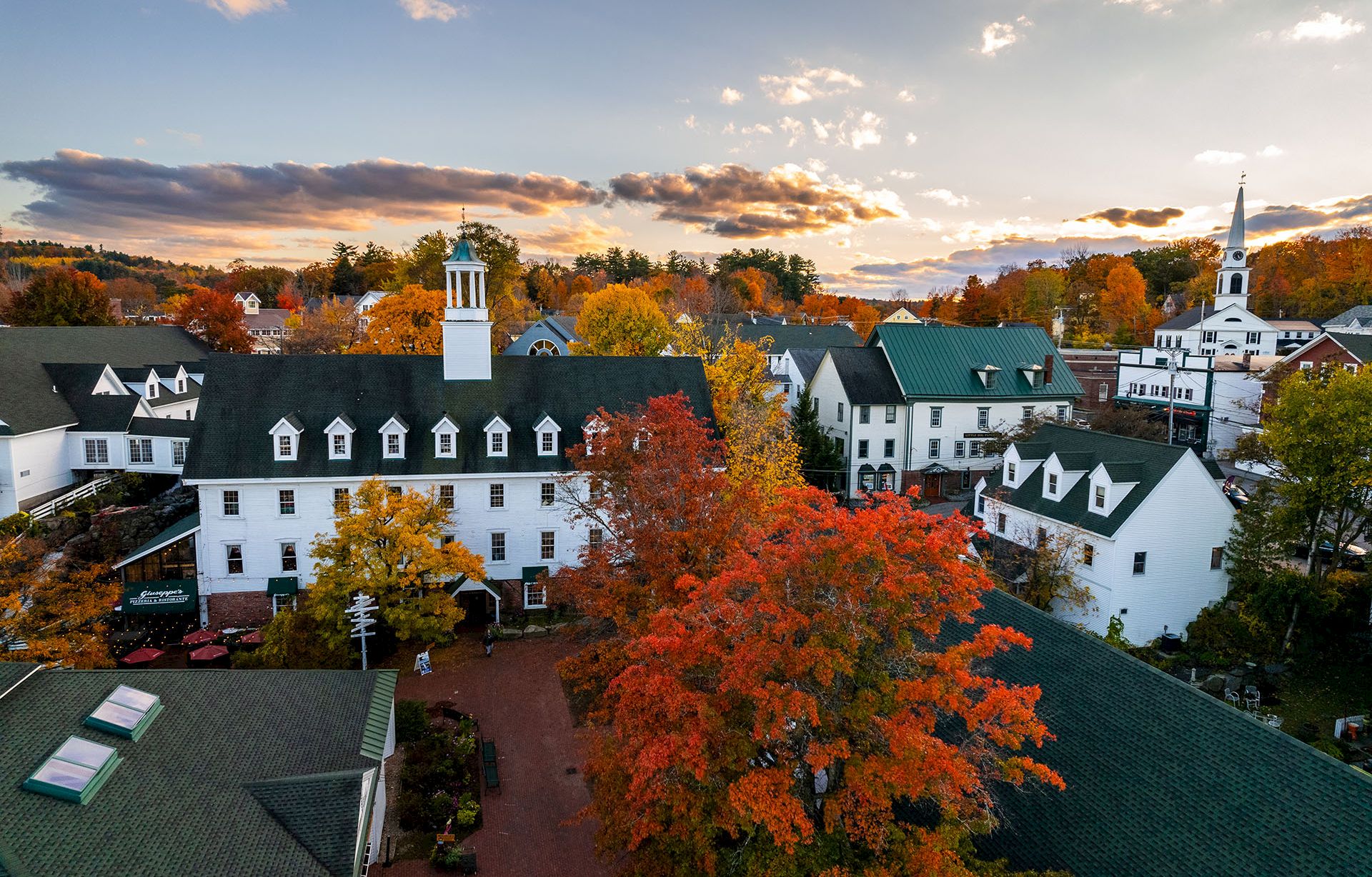 An aerial view of a white campus building with a steeple, surrounded by vivid autumn trees at sunset.