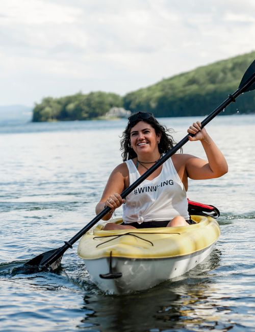 A person paddling a small kayak on calm water, wearing a life jacket and smiling as they row with a double-bladed paddle. The shoreline and trees are in the background.