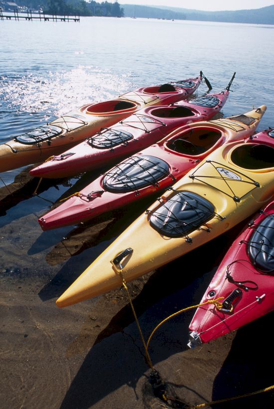 Colorful kayaks lined up at the water&rsquo;s edge, tethered to a dock on a sunny, calm lake.