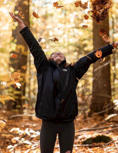 In an autumn forest, a woman throws leaves into the air with arms out, smiling as sunlight glows through the trees.