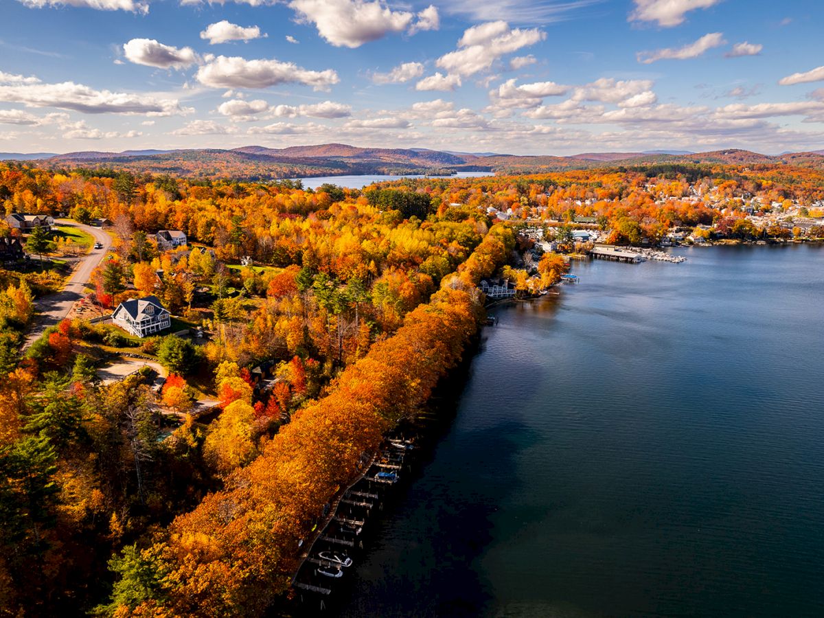 Aerial view of an autumn landscape with a lakeshore, colorful trees, houses, and a winding road under a blue sky with scattered clouds.