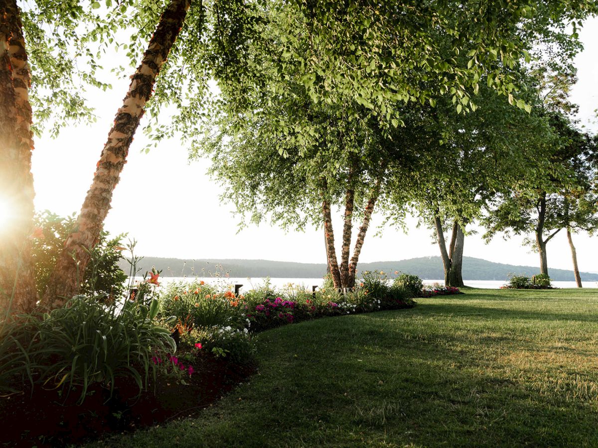 A sunny lakeside park with green lawn, trees, and flowers along the shore as the sun shines through the trees.