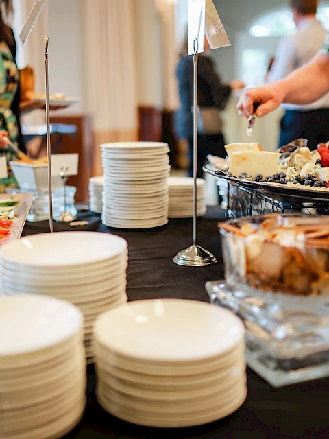 A buffet scene with stacked plates, a variety of salads and pastries, and guests serving themselves at a table.