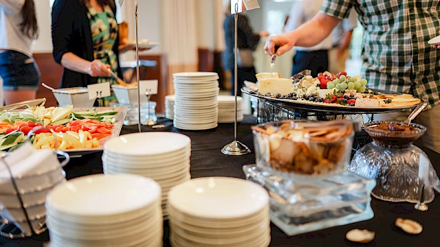 A buffet scene with stacked plates, a variety of salads and pastries, and guests serving themselves at a table.