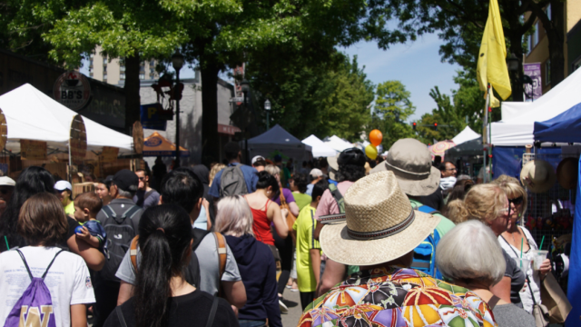 A bustling outdoor market with many people walking among stalls and booths, under tall green trees, in a cheerful and vibrant atmosphere.