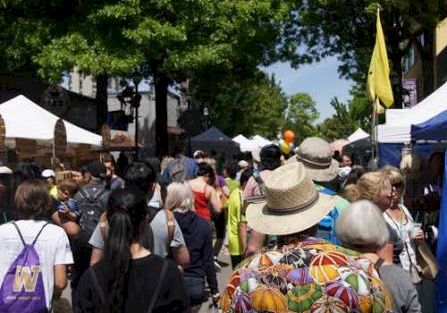 A bustling outdoor festival with tents and a crowd of people strolling under green trees.