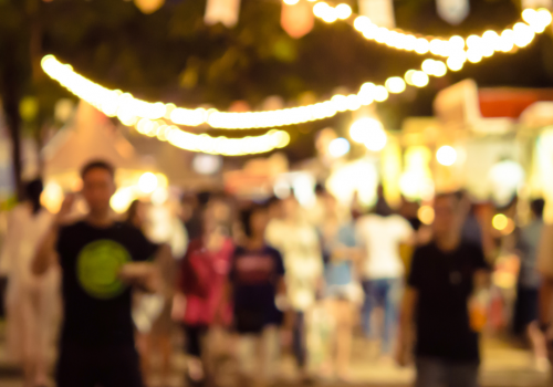 A blurred image of a festive, outdoor night market with string lights, colorful banners, and crowds of people in the background with wooden planks in the foreground.