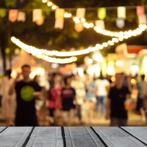 A blurred image of a festive, outdoor night market with string lights, colorful banners, and crowds of people in the background with wooden planks in the foreground.