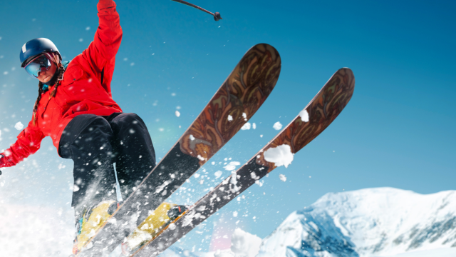 A person in a red jacket and helmet is skiing down a snowy slope, creating a spray of snow with mountains in the background.