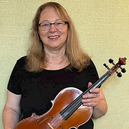 A person with long hair is smiling, holding a violin, standing against a light-colored background.