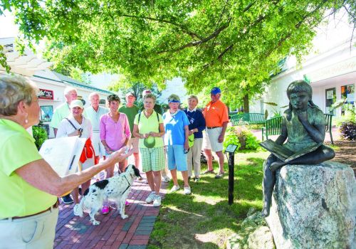 Tour group listening to a tour guide near a seated statue in a sunny park.