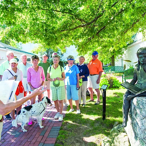 Tour group listening to a tour guide near a seated statue in a sunny park.