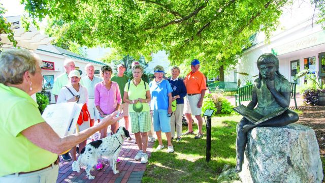 Tour group listening to a tour guide near a seated statue in a sunny park.