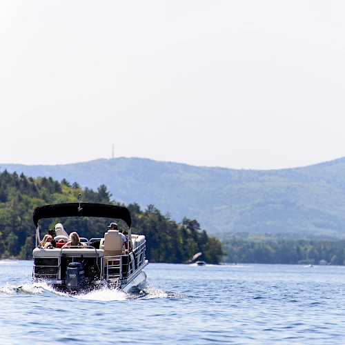 A small motorboat on a lake with people onboard, water, and distant tree-lined hills/mountains under a clear sky. Top it at 140 characters, always ending the sentence.