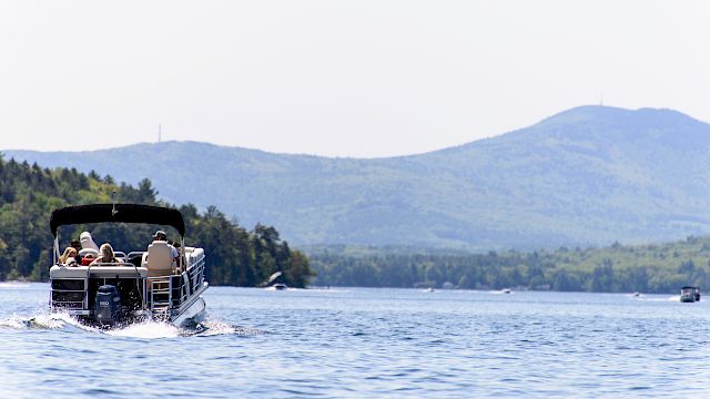 A small motorboat on a lake with people onboard, water, and distant tree-lined hills/mountains under a clear sky. Top it at 140 characters, always ending the sentence.