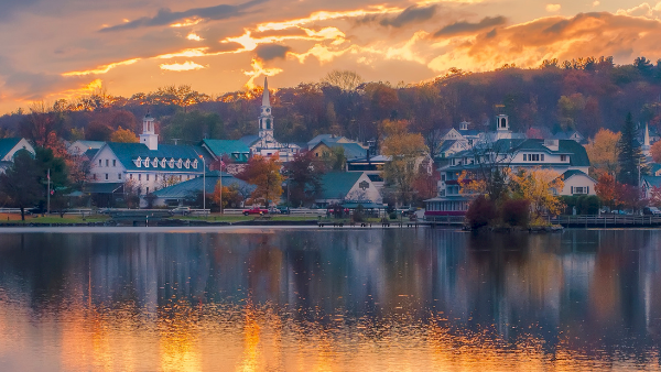 A peaceful riverside town at sunrise or sunset, with church steeples and colorful buildings reflected in a calm lake.