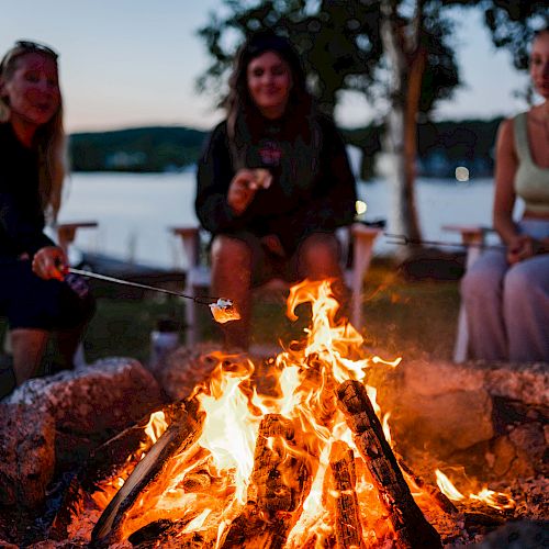 Three friends sit around a glowing campfire by the water, toasting marshmallows as dusk settles and the night approaches.