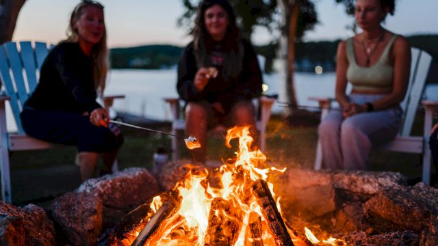 Three friends sit around a glowing campfire by the water, toasting marshmallows as dusk settles and the night approaches.