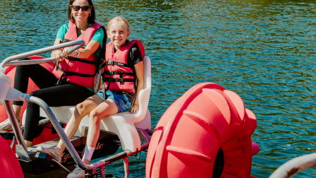 Two people wearing life jackets sit on a red pedal boat on a sunny lake, smiling as they float on the water!