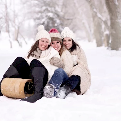 Three people in winter clothing are sitting together on the snow with a wooden sled nearby, smiling and enjoying the snowy environment.