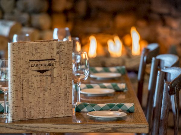 A cozy restaurant table is set with glasses, plates, and green-checkered napkins, close to a warm fireplace and a menu titled "Lakehouse."