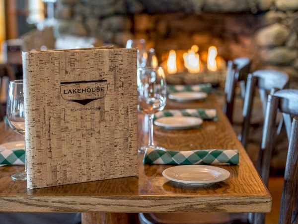 A cozy restaurant table set with a birch menu, wine glasses, rolled green-and-white napkins, plates, and a warm fireplace in the background, inviting.