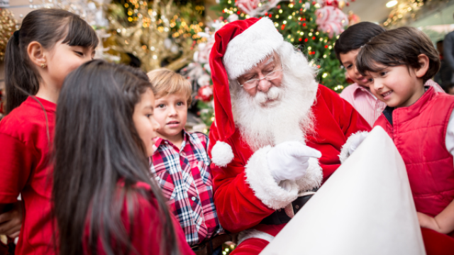 A group of children gather around Santa Claus, who is seated and holding a long scroll, in a festive Christmas setting with a decorated tree.
