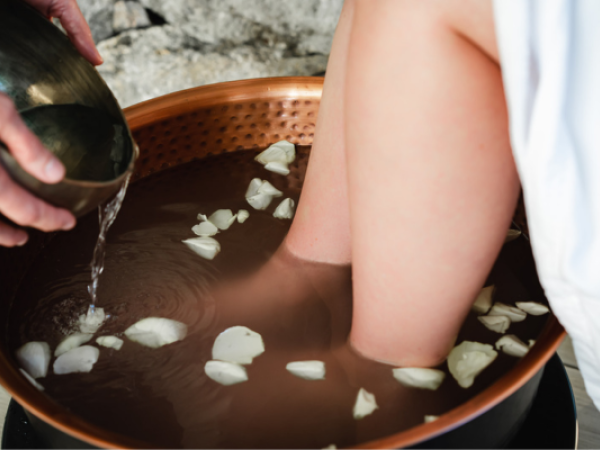 Feet soaking in a copper basin with water and rose petals, while additional water is being poured in.