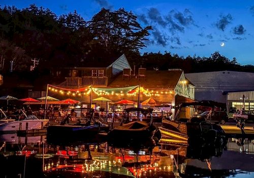 A lively waterfront scene at dusk with string lights, umbrellas, boats, and a crescent moon in the sky, reflecting on the water.