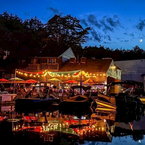 A lively waterfront scene at dusk with string lights, umbrellas, boats, and a crescent moon in the sky, reflecting on the water.