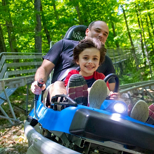 A smiling child rides a blue alpine coaster cart with a man behind him, zooming through a leafy forest trail. Adventure fun in the woods.