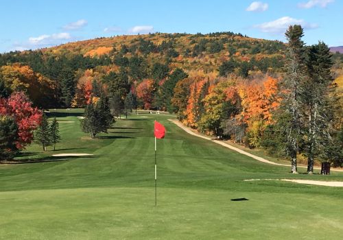A scenic golf course fairway with autumn trees, a red flag on the green, rolling hills in the background, and clear blue skies.