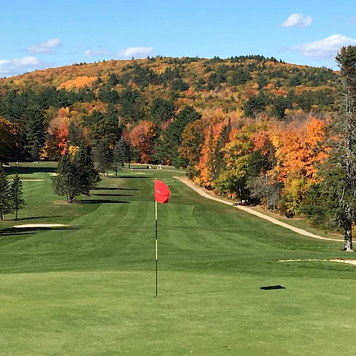A scenic golf course fairway with autumn trees, a red flag on the green, rolling hills in the background, and clear blue skies.