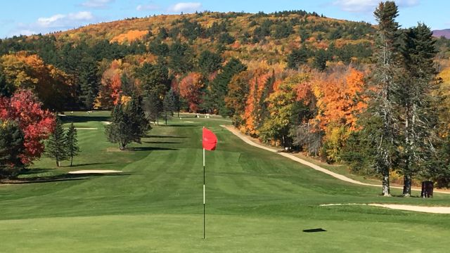 A scenic golf course fairway with autumn trees, a red flag on the green, rolling hills in the background, and clear blue skies.