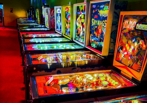 Arcade with neon-lit pinball machines in a dark hallway, colorful backlit displays, reflections on glass, vibrant game art lining the walls.