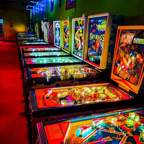 Arcade with neon-lit pinball machines in a dark hallway, colorful backlit displays, reflections on glass, vibrant game art lining the walls.