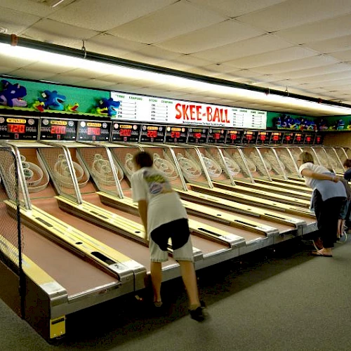 People playing skee-ball in an arcade lined with multiple alleys and a colorful sign above the lanes.