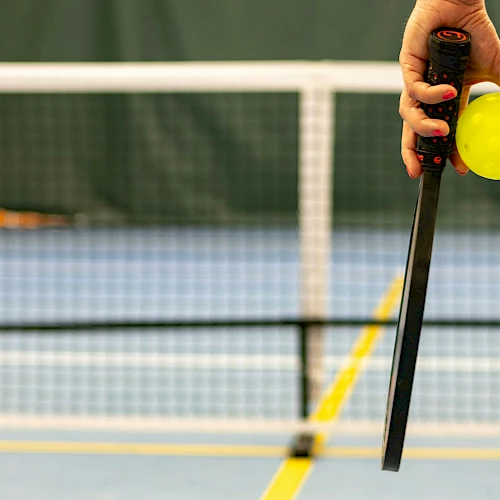 A hand holding a pickleball paddle and a yellow pickleball on an indoor court with a net in the background.