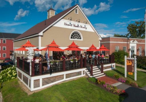 A cozy restaurant with a wooden deck, red umbrellas, outdoor seating, and a sign that reads &ldquo;Harvard Wood &hellip;&rdquo; beside a brick building.