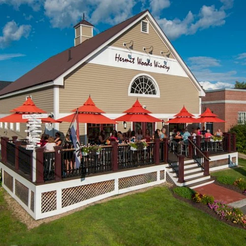 A cozy restaurant with a wooden deck, red umbrellas, outdoor seating, and a sign that reads &ldquo;Harvard Wood &hellip;&rdquo; beside a brick building.