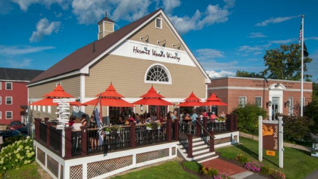 A cozy restaurant with a wooden deck, red umbrellas, outdoor seating, and a sign that reads &ldquo;Harvard Wood &hellip;&rdquo; beside a brick building.
