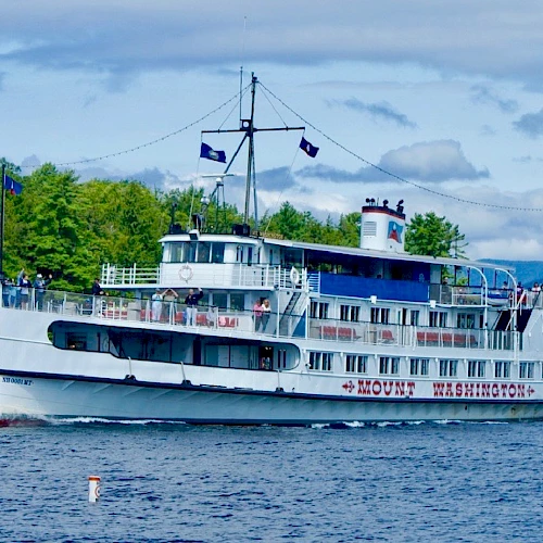 A white boat named Mount Washington cruises on a lake, with a U.S. flag atop. Tree-lined shore and hills are visible in the background.