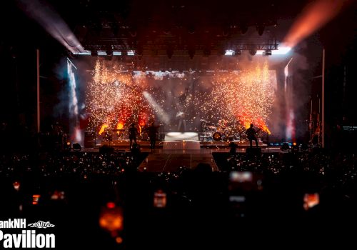 A concert stage with vibrant lighting and sparkling pyrotechnics at BankNH Pavilion, viewed by an audience holding up phones.