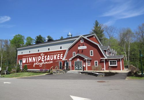 A red building labeled "WinniPesaukee Playhouse" set in a wooded area under a clear blue sky, with a road and parking area in front.
