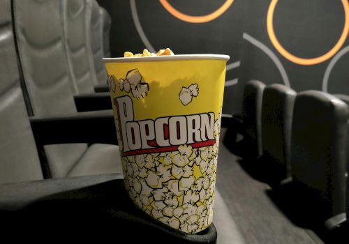 A bucket of popcorn on a theater seat, with a dark, circular pattern wall in the background.