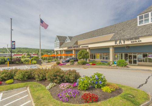 A shopping plaza with stores, a parking lot, manicured flower beds, a flagpole with the American flag, and clear weather.