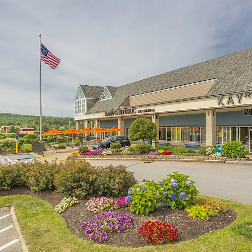 A shopping plaza with stores, a parking lot, manicured flower beds, a flagpole with the American flag, and clear weather.
