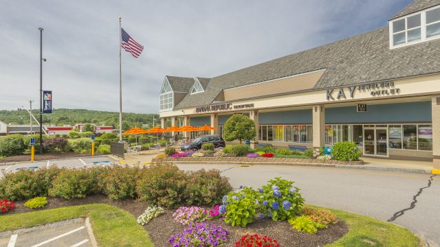 A shopping plaza with stores, a parking lot, manicured flower beds, a flagpole with the American flag, and clear weather.