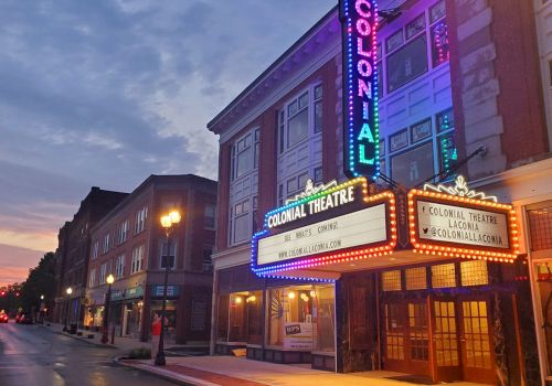 A neon-lit theater marquee on a street with brick buildings at dusk, showing &ldquo;Colonial Theatre&rdquo; signs and warm streetlights.